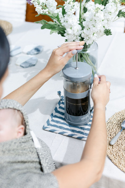 woman-with-dark-hair-pouring-coffee-from-french-press-into-mug-with-baby-asleep-in-sling-bon-and-bear